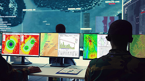 Two analysts seen from behind monitoring multiple computer screens displaying radar maps, geographic data, and surveillance visuals in a high-tech operations control room.