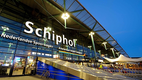 Illuminated entrance of Amsterdam Schiphol Airport at dusk, showing glass facade and signage.