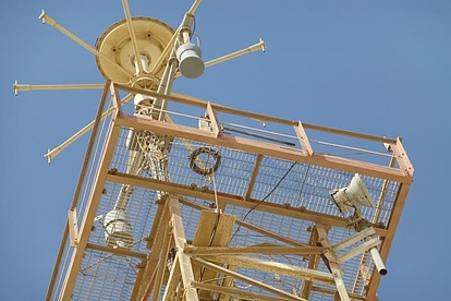 A tall communication or radar tower with metal framework and various antennas and equipment mounted on it, set against a clear blue sky.